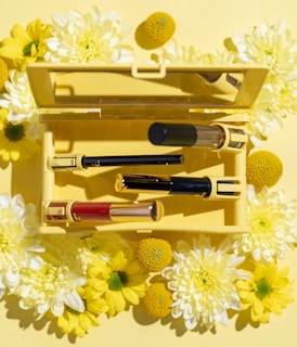 An open yellow makeup case surrounded by vibrant yellow and white flowers contains three cosmetic items. The items include tubes in black, red, and gold with sleek designs, arranged neatly inside the case.