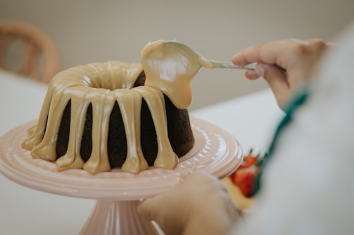 A person is icing a chocolate Bundt cake with a thick layer of creamy glaze using a spatula. The cake is on a pink cake stand, and in the background, there are blurred strawberries.