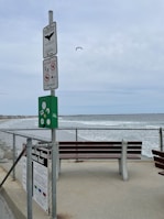 A coastal scene featuring two benches on a concrete path by the ocean, with a chain-link fence separating the path from the beach. The water appears slightly rough, and the sky is overcast. Several signs are mounted on a pole, warning about environmental and safety notices. A seagull is flying in the sky.