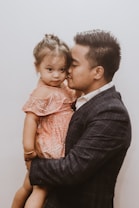 A man wearing a dark checkered blazer is holding a young girl dressed in a sparkly peach-colored dress. The man is giving the girl a kiss on the cheek while she looks towards the camera with a neutral expression. Their close interaction suggests a familial bond.