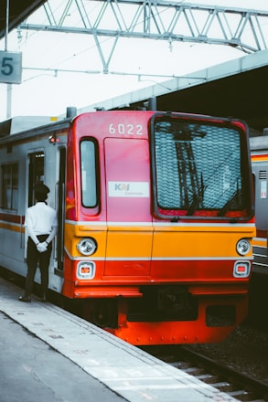 A commuter train painted in vibrant red and yellow is stationed at a platform. A person in a white shirt and black pants stands beside the train with hands clasped behind their back. Overhead, metal structures and wires are visible, indicating the station's infrastructure. The surrounding environment is urban and industrial.