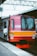 A commuter train painted in vibrant red and yellow is stationed at a platform. A person in a white shirt and black pants stands beside the train with hands clasped behind their back. Overhead, metal structures and wires are visible, indicating the station's infrastructure. The surrounding environment is urban and industrial.