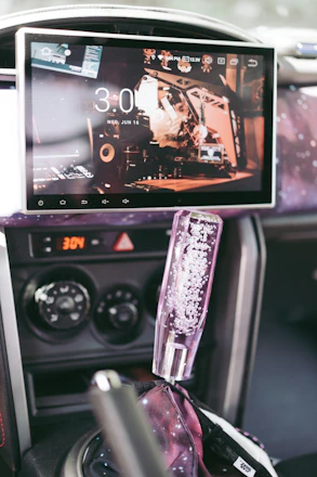 A car's interior features a large touchscreen displaying the time and a digital interface with multiple icons. The dashboard includes control knobs for air conditioning, and the gear shift has a decorative cover filled with bubbles. The surroundings have a cosmic-themed aesthetic with patterns resembling stars and galaxies.