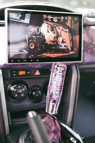 A car's interior features a large touchscreen displaying the time and a digital interface with multiple icons. The dashboard includes control knobs for air conditioning, and the gear shift has a decorative cover filled with bubbles. The surroundings have a cosmic-themed aesthetic with patterns resembling stars and galaxies.