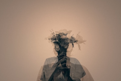 A double exposure photograph combines the silhouette of a person with a bouquet of flowers, creating a surreal and artistic effect. The person's outline is partially transparent, allowing the floral elements to stand out.