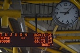 An analog clock with black numbers and hands on a white background is mounted on a ceiling structure. The clock reads approximately 10:10. Near the clock, there is an electronic display board showing red LED text with information, including destinations and times. The structure's beams are painted yellow, and the ceiling has a metallic, industrial design.