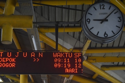 An analog clock with black numbers and hands on a white background is mounted on a ceiling structure. The clock reads approximately 10:10. Near the clock, there is an electronic display board showing red LED text with information, including destinations and times. The structure's beams are painted yellow, and the ceiling has a metallic, industrial design.