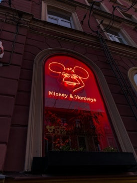 A neon red sign featuring a cartoonish face with large ears, located on a brick building facade. The sign reads 'Mickey & Monkeys' and is set within an arched window, with a small flower box below it.