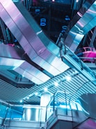 Futuristic interior architecture featuring sleek metallic surfaces and geometric patterns with vibrant blue and pink lighting. The space is dominated by large reflective panels and intricate structural elements, creating a modern, sci-fi ambiance.