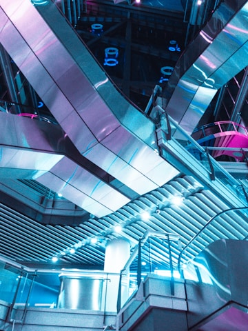 Futuristic interior architecture featuring sleek metallic surfaces and geometric patterns with vibrant blue and pink lighting. The space is dominated by large reflective panels and intricate structural elements, creating a modern, sci-fi ambiance.