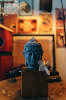 A serene blue Buddha head sculpted with closed eyes is placed on a wooden block. A delicate white flower with a yellow center rests gently against the sculpture. Wisps of smoke rise gracefully from the top, suggesting the presence of incense. The background features a variety of colorful and artistic posters, contributing to a warm, eclectic atmosphere.