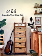 The interior of a cozy coffee shop featuring a wooden cabinet with wicker drawers, a potted plant, and a guitar resting against the wall. On the top of the cabinet, there is a small vase with dried flowers and a decorative item. The wall has the text 'Aree Coffee Slow Bar' along with Thai characters. To the right, there is a wooden counter with a laptop, some books, and art frames. Shelves on the wall hold coffee-related items like a kettle and coffee grinder.