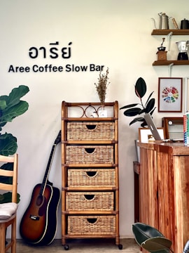 The interior of a cozy coffee shop featuring a wooden cabinet with wicker drawers, a potted plant, and a guitar resting against the wall. On the top of the cabinet, there is a small vase with dried flowers and a decorative item. The wall has the text 'Aree Coffee Slow Bar' along with Thai characters. To the right, there is a wooden counter with a laptop, some books, and art frames. Shelves on the wall hold coffee-related items like a kettle and coffee grinder.