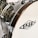 Close-up view of a drum with a wooden rim and chrome hardware. The surface is glossy with a textured pattern, and a logo is visible on the drumhead.