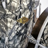 A close-up of a drum with a shiny, metallic surface and a prominent vintage-style badge labeled 'Origine 1921.' The textured material has a crumpled metal appearance. Parts of the drum hardware, such as tension rods and lugs, can be seen.
