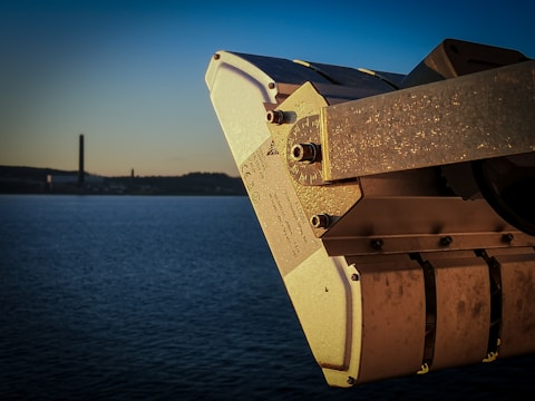 A close-up of a metallic industrial device with bolts and markings. The background features a calm body of water and a silhouette of distant industrial structures against a gradient sky transitioning from blue to a warm golden hue.