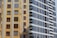 Tall residential buildings with multiple windows and balconies, showcasing architectural differences in colors and styles. One building has a yellow and beige facade with brown window frames, while the other is gray with white accents and modern design.