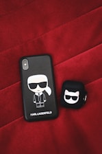 A black phone case and a black earbud case with cartoon illustrations are placed on a red textured surface. The phone case features a cartoon character with sunglasses and a white ponytail with the name 'Karl Lagerfeld' inscribed beneath. The earbud case has a cartoon cat wearing sunglasses.