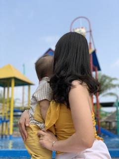 A woman with long dark hair is holding a baby and facing away from the camera. They are outdoors in a playground setting with bright playground equipment visible in the background, such as slides and ladders. The woman is wearing a yellow outfit, and the baby is dressed in a striped shirt and yellow pants.