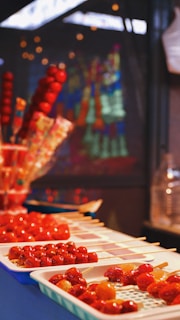 Skewers of glazed red fruit arranged on trays, with vibrant colors and shiny surfaces reflecting light. In the background, blurred colorful decorations are visible, suggesting an outdoor or market setting.