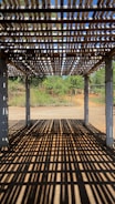 A wooden pergola casting intricate shadow patterns on the ground, with concrete pillars supporting the structure. In the background, lush greenery and a clear blue sky are visible, suggesting a serene outdoor setting.