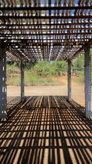 A wooden pergola casting intricate shadow patterns on the ground, with concrete pillars supporting the structure. In the background, lush greenery and a clear blue sky are visible, suggesting a serene outdoor setting.