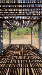 A wooden pergola casting intricate shadow patterns on the ground, with concrete pillars supporting the structure. In the background, lush greenery and a clear blue sky are visible, suggesting a serene outdoor setting.