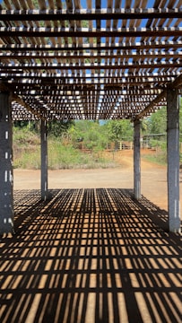 A wooden pergola casting intricate shadow patterns on the ground, with concrete pillars supporting the structure. In the background, lush greenery and a clear blue sky are visible, suggesting a serene outdoor setting.