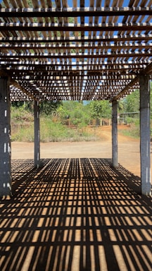 A wooden pergola casting intricate shadow patterns on the ground, with concrete pillars supporting the structure. In the background, lush greenery and a clear blue sky are visible, suggesting a serene outdoor setting.