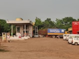 A rural gas station with several fuel pumps and a small building with signage in another language. There is a large truck with a blue container labeled 'Nazrul Transport Agency' parked nearby. Several white vans and cars are also visible, along with a bright red building labeled 'Shahser Ali Hotel and Restaurant'. Trees and greenery surround the area, and a ladder is visible leaning against the building.