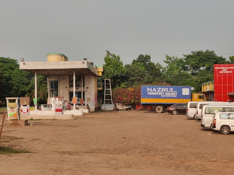 A rural gas station with several fuel pumps and a small building with signage in another language. There is a large truck with a blue container labeled 'Nazrul Transport Agency' parked nearby. Several white vans and cars are also visible, along with a bright red building labeled 'Shahser Ali Hotel and Restaurant'. Trees and greenery surround the area, and a ladder is visible leaning against the building.