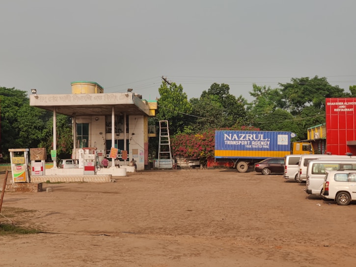 A rural gas station with several fuel pumps and a small building with signage in another language. There is a large truck with a blue container labeled 'Nazrul Transport Agency' parked nearby. Several white vans and cars are also visible, along with a bright red building labeled 'Shahser Ali Hotel and Restaurant'. Trees and greenery surround the area, and a ladder is visible leaning against the building.