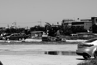An excavator is situated in an open area with several parked cars nearby. The setting appears to be on the outskirts of a city with buildings, utility poles, and some vegetation in the background. The overall environment looks industrial and somewhat isolated, with a dirt or gravel lot.