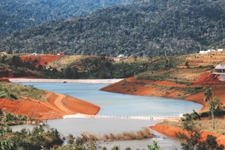 A picturesque landscape featuring a winding river surrounded by lush greenery and red clay soil. The terrain includes rolling hills and dense forested areas in the background. Small structures and possibly a dam or barrier are visible, indicating some human alteration of the landscape.