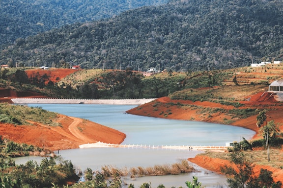 A picturesque landscape featuring a winding river surrounded by lush greenery and red clay soil. The terrain includes rolling hills and dense forested areas in the background. Small structures and possibly a dam or barrier are visible, indicating some human alteration of the landscape.
