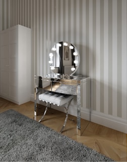 A stylish vanity area featuring a reflective, mirrored dressing table with a round mirror framed by round lights. In front of the table is a padded stool with a tufted design. The room includes a striped wallpaper, a soft gray rug, and light wood flooring.