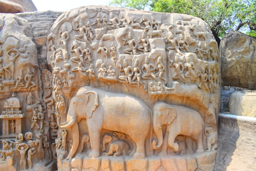 A large stone carving depicting elephants, numerous human figures, and animals intricately formed as part of an ancient mural. The artwork is set on a rocky surface with natural textures, showcasing detailed sculptures of people in various poses, possibly representing a narrative or mythological scene. The setting is outdoors with some greenery visible at the top.