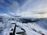 An expansive view of snow-covered mountains under a dramatic, cloud-filled sky. In the foreground, there are structures with antennas and equipment, likely a weather station, perched on the mountain ridge.