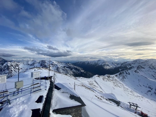 An expansive view of snow-covered mountains under a dramatic, cloud-filled sky. In the foreground, there are structures with antennas and equipment, likely a weather station, perched on the mountain ridge.