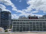 A cityscape with several modern and traditional buildings. The sky is clear with some scattered clouds. A prominent building features a large sign with Korean text.