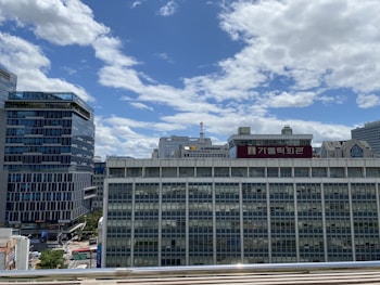 A cityscape with several modern and traditional buildings. The sky is clear with some scattered clouds. A prominent building features a large sign with Korean text.
