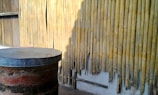 A weathered bamboo wall with visible wear and tear, casting shadows in the sunlight. In the foreground, an old, rustic barrel with a metal lid sitting on a wooden platform is visible.