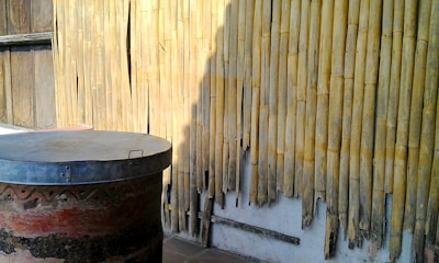 A weathered bamboo wall with visible wear and tear, casting shadows in the sunlight. In the foreground, an old, rustic barrel with a metal lid sitting on a wooden platform is visible.