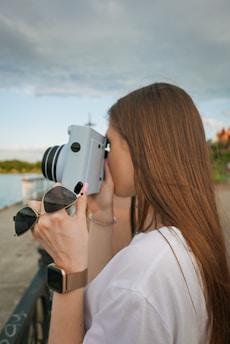 A woman with long brown hair holds a white instant camera to her eye, appearing to take a photo. In her other hand, she holds a pair of sunglasses. She wears a white shirt and a smart watch with a metallic band on her wrist. The background features a cloudy sky, with a body of water and shoreline visible, suggesting an outdoor setting.