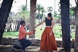 A man is kneeling down, offering his hand to a woman who is standing. They are outdoors, surrounded by palm trees and decorative architecture. The man is wearing a coral shirt with dark pants and sunglasses, while the woman is wearing a long, orange skirt, a black top, and sunglasses. The setting suggests a warm, sunny day in a park or garden area.
