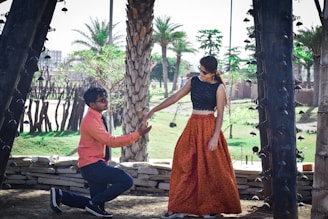 A man is kneeling down, offering his hand to a woman who is standing. They are outdoors, surrounded by palm trees and decorative architecture. The man is wearing a coral shirt with dark pants and sunglasses, while the woman is wearing a long, orange skirt, a black top, and sunglasses. The setting suggests a warm, sunny day in a park or garden area.