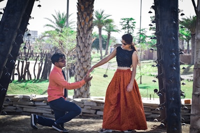 A man is kneeling down, offering his hand to a woman who is standing. They are outdoors, surrounded by palm trees and decorative architecture. The man is wearing a coral shirt with dark pants and sunglasses, while the woman is wearing a long, orange skirt, a black top, and sunglasses. The setting suggests a warm, sunny day in a park or garden area.