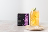 A glass filled with a refreshing beverage containing ice, lemon, and lime slices is placed on a wooden coaster beside a black and purple box labeled 'Elixir Detox AGATA' from Crystaltea. The setup is against a light, minimalist background.