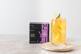 A glass filled with a refreshing beverage containing ice, lemon, and lime slices is placed on a wooden coaster beside a black and purple box labeled 'Elixir Detox AGATA' from Crystaltea. The setup is against a light, minimalist background.