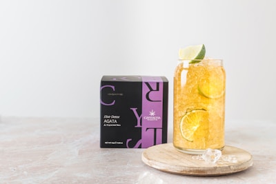 A glass filled with a refreshing beverage containing ice, lemon, and lime slices is placed on a wooden coaster beside a black and purple box labeled 'Elixir Detox AGATA' from Crystaltea. The setup is against a light, minimalist background.